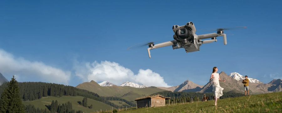 Drone flying over a scenic landscape with mountains and a wooden cabin. Sold by an Authorized DJI Retailer.