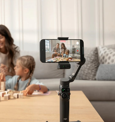 Smartphone on a DJI gimbal stabilizer with a screen displaying a video call, placed on a table with a family in the background. Sold by a DJI Authorized Retailer.